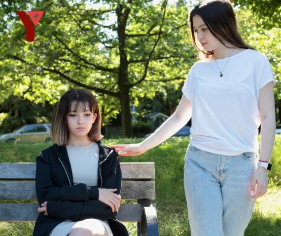 A teen girl on a bench looking sad, being comforted by a friend standing next to her.